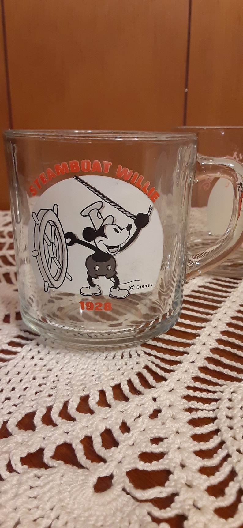 Puede incluir: Una taza de vidrio transparente con una imagen en blanco y negro de Mickey Mouse como Steamboat Willie. La taza tiene el texto "Steamboat Willie" y "1928" en letras naranjas. La taza tambi&eacute;n tiene el s&iacute;mbolo de copyright de Disney.