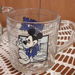 Puede incluir: Una taza de vidrio transparente con un dise&ntilde;o de Mickey Mouse azul y blanco. Mickey lleva un traje azul y sostiene un trozo de papel. La taza tiene una franja azul alrededor de la parte superior y un asa.