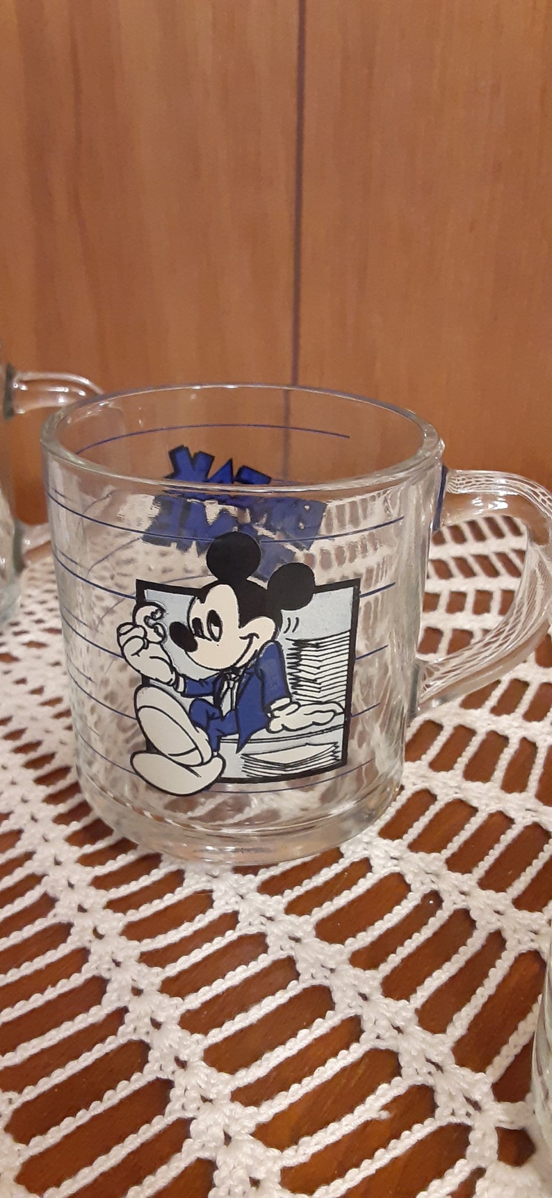 Puede incluir: Una taza de vidrio transparente con un dise&ntilde;o de Mickey Mouse azul y blanco. Mickey lleva un traje azul y sostiene un trozo de papel. La taza tiene una franja azul alrededor de la parte superior y un asa.