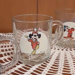 Puede incluir: Dos tazas de vidrio transparente con asas. La taza de la izquierda tiene un logotipo de Mickey Mouse Club con Mickey Mouse tocando la trompeta. La taza de la derecha tiene un logotipo de Mickey Mouse Club con Mickey Mouse usando un sombrero de mago. Ambas tazas tienen el texto "Mickey Mouse Club" y "Disney".