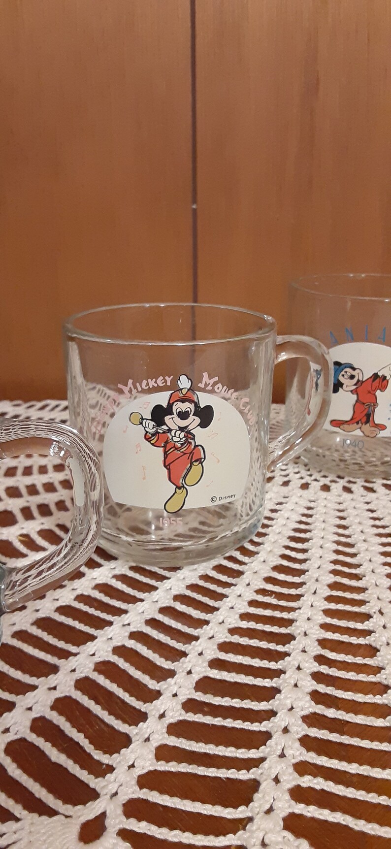 Puede incluir: Dos tazas de vidrio transparente con asas. La taza de la izquierda tiene un logotipo de Mickey Mouse Club con Mickey Mouse tocando la trompeta. La taza de la derecha tiene un logotipo de Mickey Mouse Club con Mickey Mouse usando un sombrero de mago. Ambas tazas tienen el texto "Mickey Mouse Club" y "Disney".