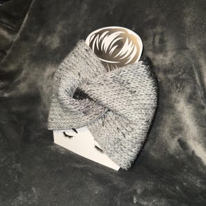 May include: A gray knit headband with a twisted knot design. The headband has black flecks throughout and is displayed on a white card with a gold logo and a pair of false eyelashes.