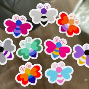 May include: A collection of colorful butterfly stickers with various designs. Each sticker features a butterfly shape with different color schemes, including pink, purple, blue, orange, and green. The stickers have a white border and are likely for decoration or crafting.
