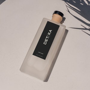 May include: A clear glass bottle with a black label that says "DETKA" and "VETIVER" on a white surface. The bottle has a black cap.