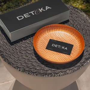 May include: A copper serving bowl with a hammered finish sits on a black woven surface. A black business card with the text "DET&KA" is inside the bowl.