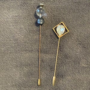 Puede incluir: Dos alfileres decorativos para sombreros. Uno presenta un globo de cristal transparente y una cuenta gris oscuro. El otro tiene un marco cuadrado dorado con una gema clara y facetada. Ambos alfileres tienen largos y delgados vástagos metálicos.