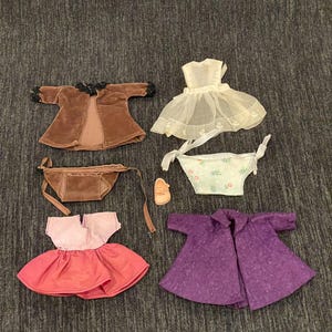 May include: A collection of vintage doll clothes, including a brown velvet coat with black trim, a white dress, a floral-print diaper, a pink and red dress, a purple coat, a brown purse, and a small shoe.