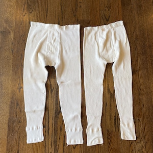 Puede incluir: Dos pares de pantalones de ropa interior térmica de color blanco roto. Los pantalones son acanalados y tienen bragueta abotonada y puños acanalados en los tobillos. Las prendas están sobre una superficie de madera oscura.