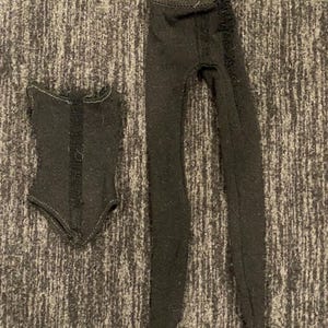 May include: A black leotard and black tights for a doll. The leotard has a ruffled neckline and a seam down the front. The tights are plain.