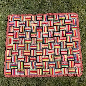 May include: A patchwork quilt with a red, ruffled border. The quilt features numerous rectangular blocks in various colours, including red, yellow, blue, and green. The quilt is placed on green grass.