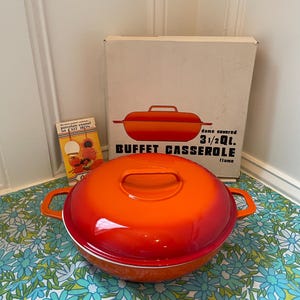 Puede incluir: Una fuente para bufé de color naranja, con tapa y asas. El plato está sobre una superficie con estampado floral. La caja detrás del plato dice "BUFFET CASSEROLE" y "3 1/2 Qt."