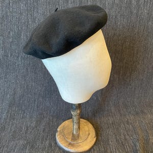 May include: A black beret hat displayed on a mannequin head. The beret is made of a soft, dark material and has a classic, round shape. The mannequin head is a light beige color and is mounted on a wooden stand.