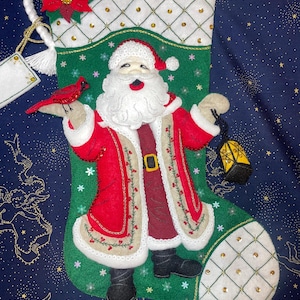 May include: A felt Christmas stocking with a green background and a white, red, and beige Santa Claus holding a lantern. The stocking is decorated with gold glitter and rhinestones.