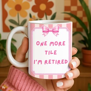 May include: A white ceramic mug featuring a pink and white vertical striped background with a pink bow at the top. The centre contains a decorative frame with the pink text: ONE MORE TILE I'M RETIRED. The mug has a curved handle on the left side.