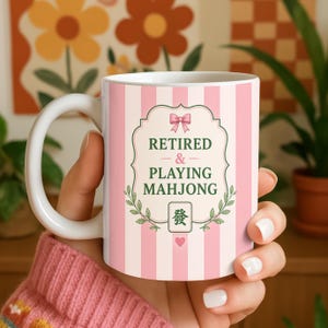 Könnte beinhalten: Weißer Keramikbecher mit rosa vertikalen Streifen. Der Becher zeigt den Text "RETIRED & PLAYING MAHJONG" in einem dekorativen Rahmen mit einer Schleife und floralen Akzenten. Ein chinesisches Schriftzeichen und ein Herz sind ebenfalls enthalten.