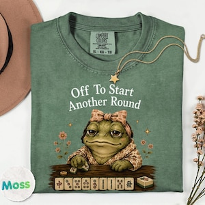 May include: A sage green t-shirt featuring a graphic of a toad wearing a patterned headpiece, sitting at a table with mahjong tiles. The text reads "Off To Start Another Round" in white lettering. The shirt has a Comfort Colors brand label.