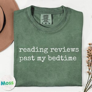 May include: A sage green crewneck t-shirt with the white text reading reviews past my bedtime printed on the front. The shirt features a Comfort Colors brand label at the inner neckline and has a short-sleeved, casual design.