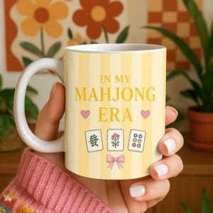 Könnte beinhalten: Weißer Keramikbecher mit gelb gestreiftem Hintergrund und dem Text "IN MY MAHJONG ERA" in Gold. Der Becher zeigt rosa Herzen und Mahjong-Kachel-Illustrationen. Der Becher wird von einer Person gehalten.