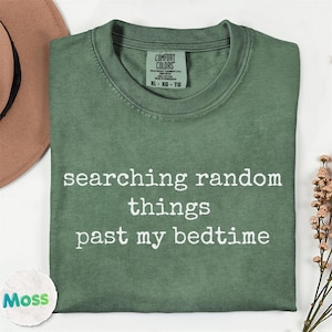 May include: A sage green crew neck t-shirt with the white text "searching random things past my bedtime" printed on the front. The shirt features a Comfort Colors brand label at the inner neckline and is made of 100% cotton fabric.