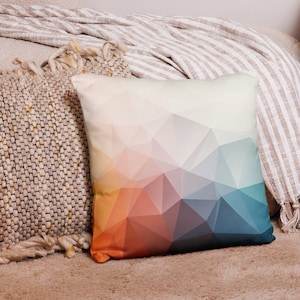 May include: A geometric patterned throw pillow with a gradient of orange, red, blue, and white colors. The pillow is on a light brown fuzzy surface.