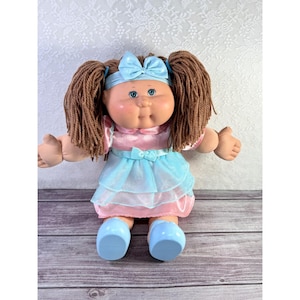 May include: A vintage Cabbage Patch Kids doll with brown yarn pigtails, blue eyes, and rosy cheeks. The doll wears a pink and blue dress with a matching blue bow and headband. Blue shoes complete the outfit. The doll is in a seated position.