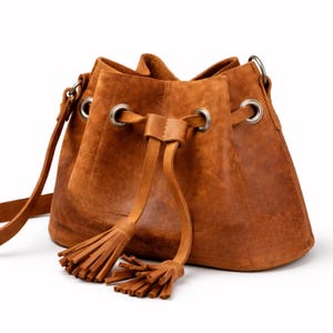 May include: A brown leather bucket bag with a drawstring closure and a shoulder strap. The bag features silver-toned metal grommets and a decorative tassel. The leather has a textured, distressed appearance.