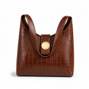 May include: A brown handbag with a crocodile-embossed texture, featuring two integrated shoulder straps and a central vertical tab closure secured by a large, circular, smooth gold-toned metal button centred on the front panel.