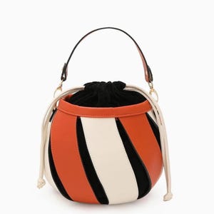 May include: A round handbag with a black handle and a drawstring closure. The bag features a color block design with orange, white, and black panels. The bag has a cream-colored drawstring and a black interior.