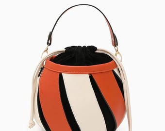 May include: A round handbag with a black handle and a drawstring closure. The bag features a color block design with orange, white, and black panels. The bag has a cream-colored drawstring and a black interior.