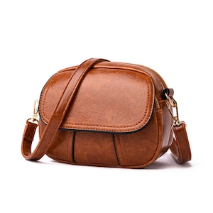May include: A brown leather crossbody bag with a textured finish. The bag has a rounded shape with a flap closure and a long adjustable strap. Gold-colored hardware accents the bag, including the zipper and strap attachments.