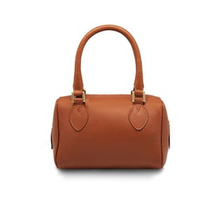 May include: A brown leather handbag with a cylindrical shape and two short handles. The bag has gold-colored hardware and a textured surface. The handles are arched and attached to the bag with gold-colored metal pieces.