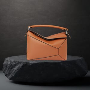 Op de afbeelding: Een bruinleren handtas met een geometrisch ontwerp, met een handvat aan de bovenkant en een schouderriem. De tas wordt getoond op een donkere, getextureerde steen, die de hoekige constructie en contrasterende stiksels benadrukt. Afmetingen: 30 cm breed, 20 cm hoog, 10 cm diep.