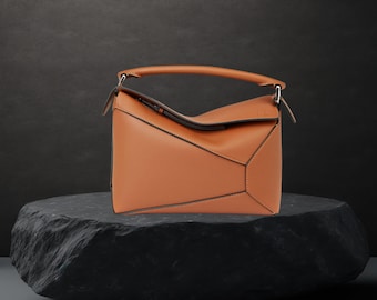 May include: A tan leather handbag with a geometric design, featuring a top handle and a shoulder strap. The bag is displayed on a dark, textured stone, showcasing its angular construction and contrasting stitching. The bag's dimensions are approximately 12 inches wide, 8 inches high, and 4 inches deep.