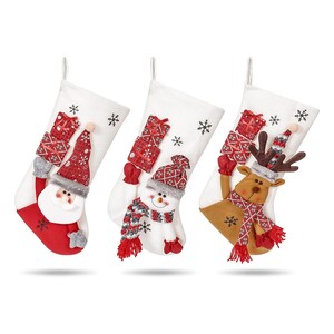 Peut inclure: Trois bas de Noël blancs avec des motifs festifs. L'un représente le Père Noël, un autre un bonhomme de neige et le troisième un renne. Chaque bas a des accents rouges, des motifs de flocons de neige et une boucle pour accrocher.