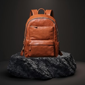 以下が含まれることがあります： 複数のジッパー付きコンパートメントと上部ハンドルを備えた茶色の革製バックパック。 バックパックは、そのデザインと職人技を示す、暗く質感のある表面に表示されています。 バッグには調節可能なショルダーストラップが付いています。