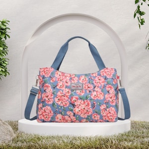 Può includere: Una borsa tote floreale con sfondo blu e fiori rosa. La borsa ha manici blu e una tracolla staccabile. Il testo "Sue Ye To" è visibile sulla parte anteriore della borsa. La borsa è appoggiata su un piedistallo bianco.