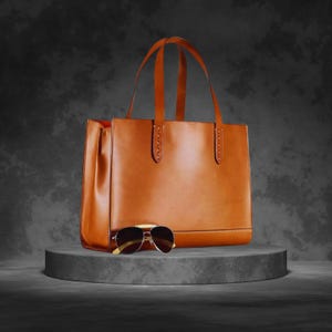 Könnte beinhalten: Eine braune Ledertasche mit zwei Henkeln und eine Sonnenbrille. Die Tasche ist rechteckig mit einem strukturierten Design. Die Sonnenbrille hat einen goldenen Rahmen und dunkle Gläser. Die Artikel werden auf einer grauen, kreisförmigen Plattform präsentiert.