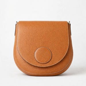 May include: A tan leather crossbody bag with a rounded flap and a circular accent on the front. The bag has a textured surface and a silver-coloured metal clasp. The stitching is a light tan colour.