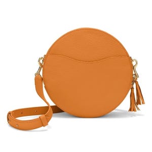 Puede incluir: Un bolso bandolera redondo naranja con una superficie texturizada. El bolso tiene una correa larga y ajustable y herrajes dorados. Un pequeño bolsillo está en la parte delantera y una borla cuelga del lateral. Un accesorio elegante.