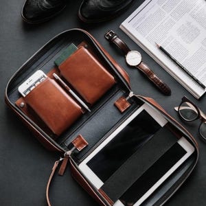 May include: A brown leather portfolio case with a zipper, open to reveal a tablet, phone, and wallet. A watch, glasses, pencil, and a magazine are arranged around the case on a black surface. The portfolio has a handle.