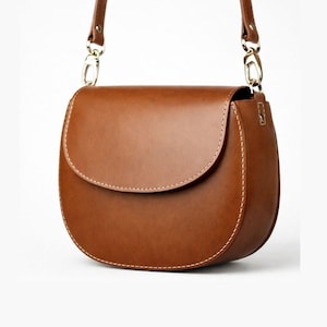 May include: A brown leather crossbody bag with a curved flap and white stitching. The bag has a long, adjustable strap and gold-colored hardware. The bag is a classic design, suitable for everyday use.