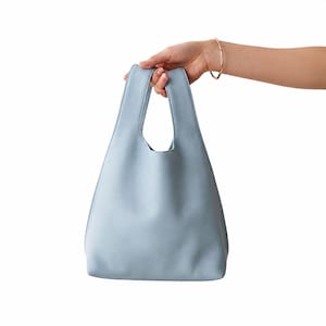 May include: A light blue tote bag with a smooth, matte finish and integrated handles. The bag features a simple, vertical, triangular silhouette with a flat base, constructed from a solid, uniform material in a soft, pale blue color.