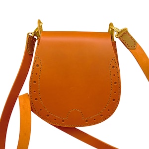 Puede incluir: Bolso bandolera estilo alforja en tono naranja cálido. El bolso tiene un cierre de solapa y una correa larga y ajustable. Las costuras decorativas y los pequeños recortes circulares adornan los bordes de la solapa. Herrajes dorados conectan la correa al bolso.