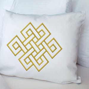 Puede incluir: Cojín cuadrado blanco con un diseño geométrico dorado bordado. El diseño es una serie de cuadrados y líneas entrelazados, creando un patrón simétrico. El cojín está sobre una superficie blanca.