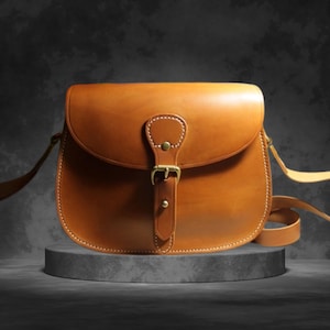 May include: A brown leather satchel bag with a curved flap and adjustable shoulder strap. The bag features a buckle closure and is displayed on a gray pedestal. The stitching is visible around the edges of the bag.
