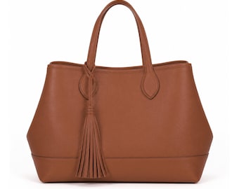 May include: A medium-brown tote bag with a structured silhouette, two top handles, and a long decorative tassel attached to one handle base. The bag features a smooth, solid-colored exterior with clean stitching along the bottom panel.
