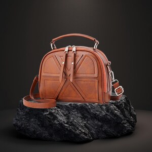 Könnte beinhalten: Eine braune, kuppelförmige Handtasche mit einem oberen Griff und einem abnehmbaren Schulterriemen. Die Tasche hat geometrische Aussparungen und silberfarbene Reißverschlüsse und Beschläge. Die Tasche steht auf einer dunklen, strukturierten Oberfläche.