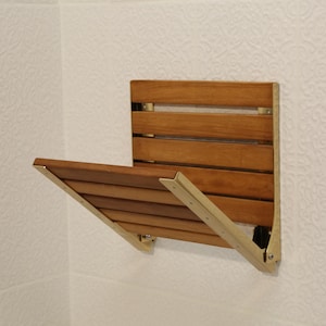 Könnte beinhalten: Ein an der Wand montierter, klappbarer Duschsitz aus Naturholz mit messingfarbenen Metallstützen. Der Sitz ist mit horizontalen Holzlatten versehen und wird bei Nichtgebrauch an die Wand geklappt. Der Sitz ist an einer strukturierten weißen Wand montiert.
