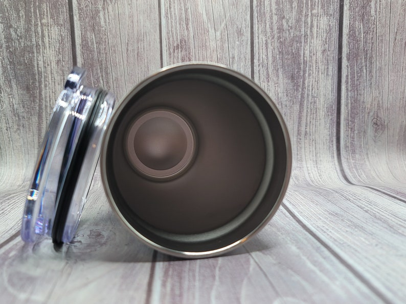 Op de afbeelding: Een roestvrijstalen tumbler met een deksel. De tumbler is zilverkleurig en heeft een zwarte rubberen ring rond de bovenkant. Het deksel is van helder plastic.