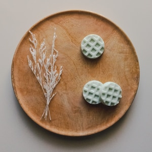 Peut inclure: Trois savons blancs en forme de gaufre sur une assiette en bois avec une branche de fleurs séchées blanches.